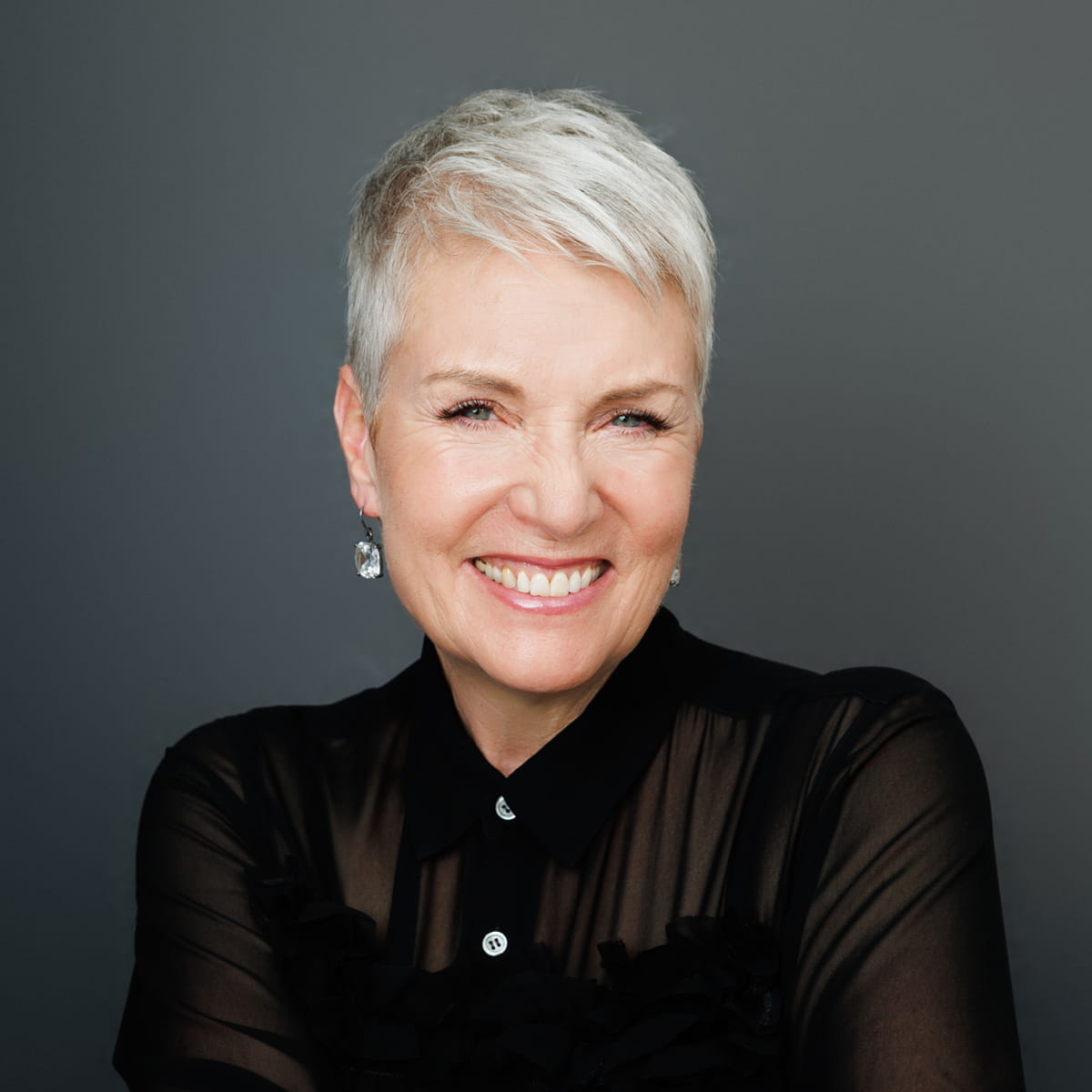 Smiling woman with short white hair wearing a sheer black top and earrings, posed against a plain gray background.