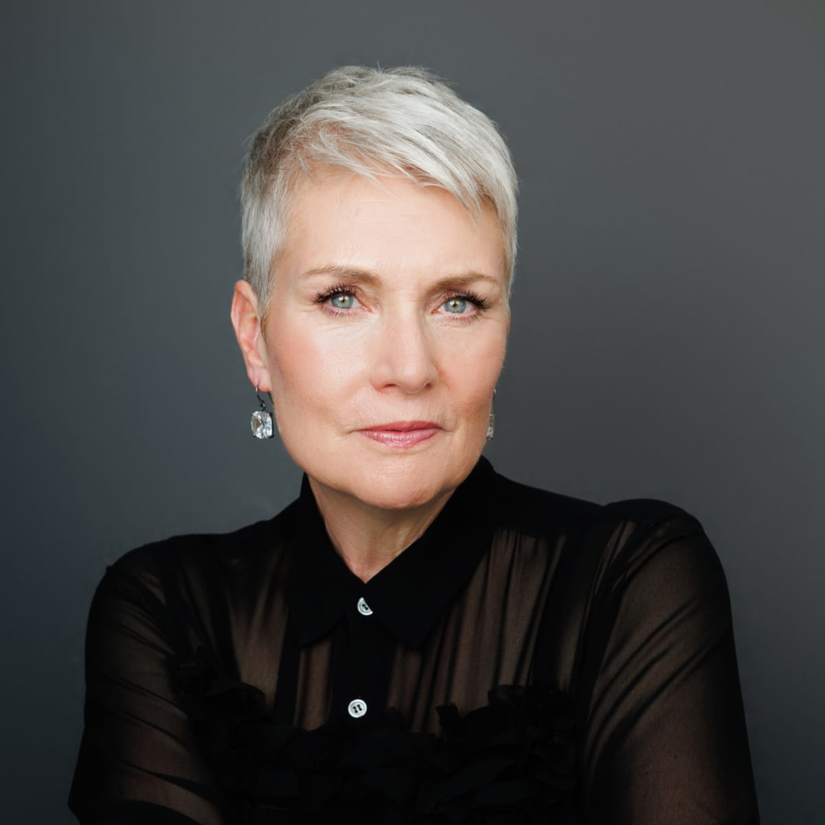 A confident woman with short white hair and light eyes, wearing a sheer black blouse and earrings, poses against a plain dark background.