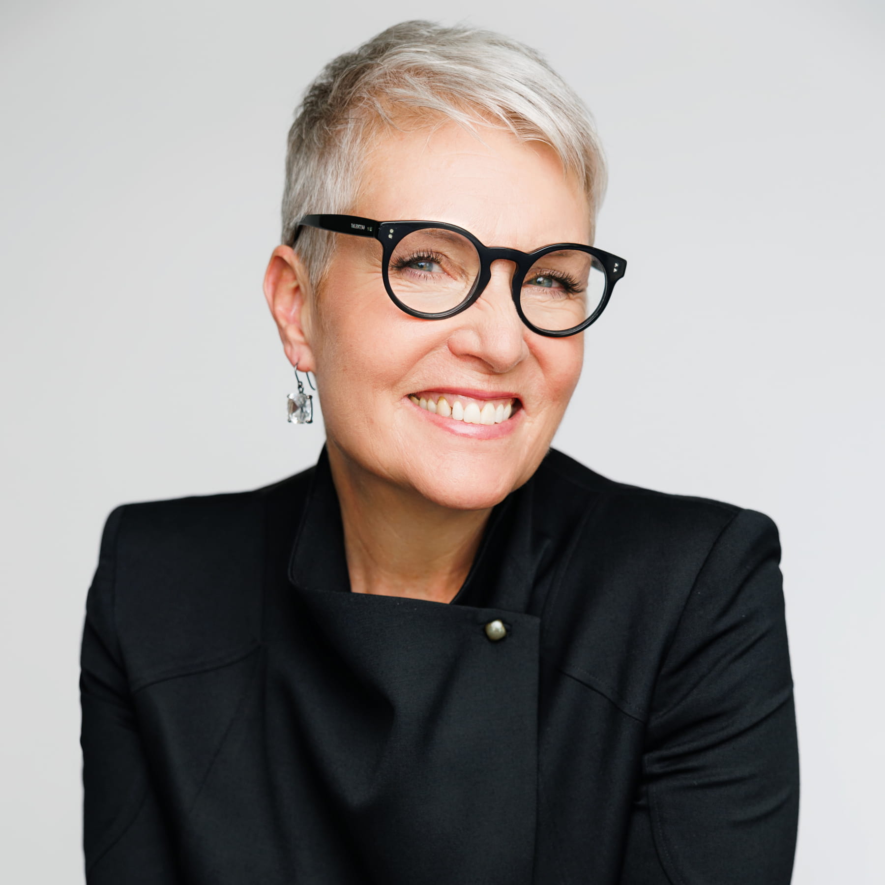 Smiling woman with short gray hair, black glasses, silver earrings, and a black jacket, posed against a plain light background.