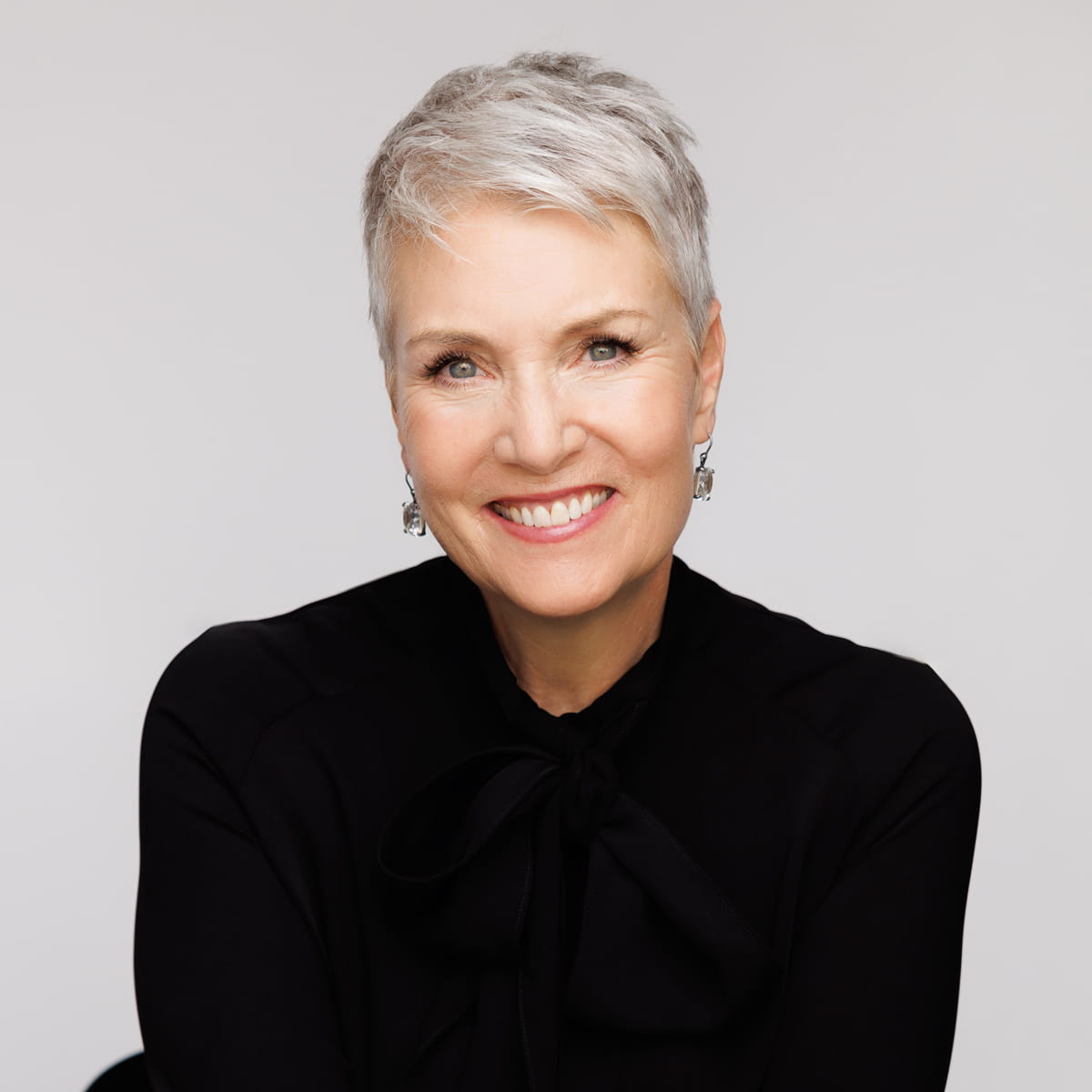 A smiling woman with short gray hair wearing a black top and silver earrings, posed against a plain light background.