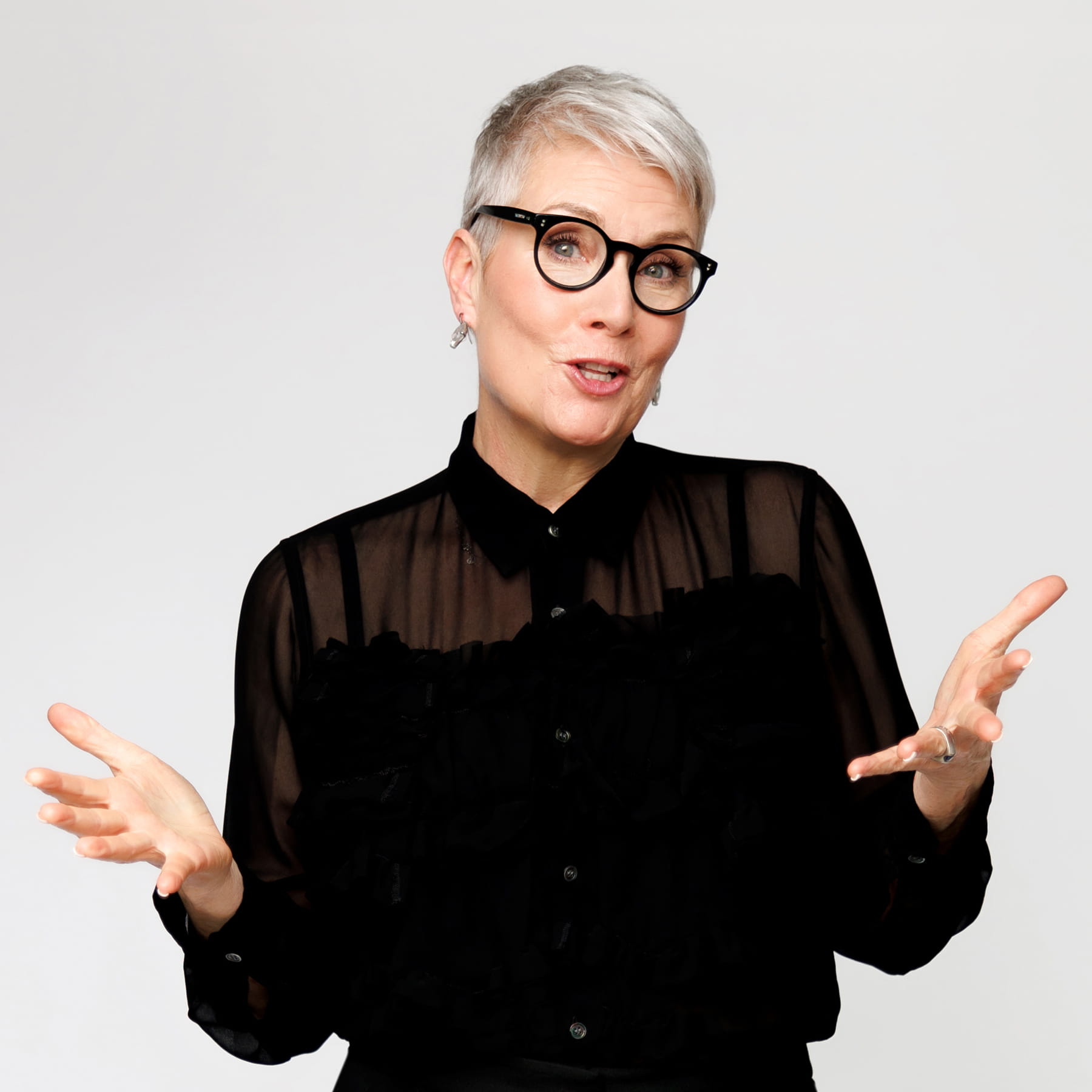 A person with short gray hair, glasses, and a sheer black blouse gestures with open hands while speaking, against a plain white background.